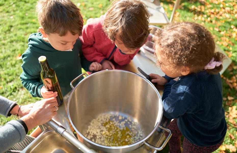Drei Kinder gucken von oben in einen großen Kochtopf.