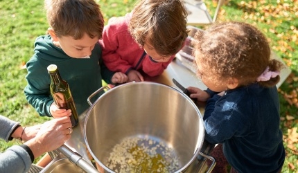 Drei Kinder gucken von oben in einen großen Kochtopf.