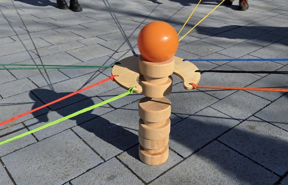 Eine Holzscheibe mit großer Einkerbung wird mit Schnüren zwischen den Teilnehmenden gespannt; Holzklötze werden so in Gemeinschaftsarbeit aufeinander gestellt