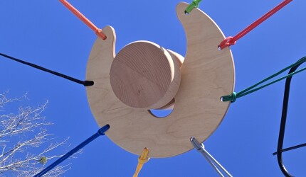 Ein Holzscheibe mit einer großen Einkerbung wird von sieben Schnüren gespannt; in de Einkerbung ist ein Holzelemet, welches von der Gruppe gemeinsam durch das Spannen der Schnüre versetzt werden muss