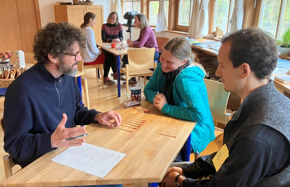 Ein Raum mit Tischen und Stühlen; je drei Teilnehmende sitzen an einem Tisch und besprechen sich; eine Gruppe mit zwei Männern und einer Frau sind im Vordergrund; im Hintergrund ein weiterer Tisch mit drei Teilnehmerinnen