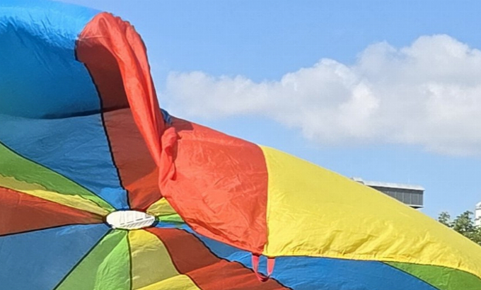 Ein buntes Schwungtuch vor einem hellblauen Himmel mit einer Wolke. Das Schwungtuch sieht einem Regenschirm ähnlich - die Farben sind blau, rot, gelb und grün