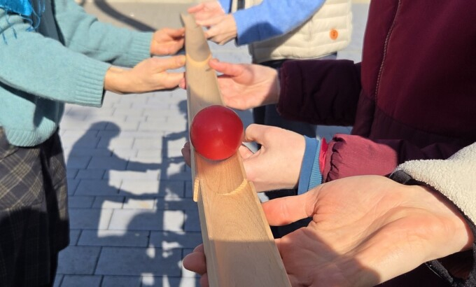 Vier Personen, von denen nur die Hände zu sehen sind, halten jeweils eine Holzrinne; ein roter Ball rollt darüber; Ziel ist es, den Ball mit den vier Holzrinnen über eine lange Distanz zu befödern