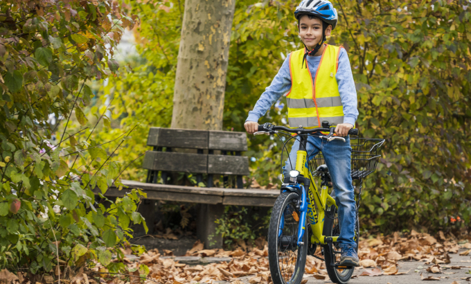 Ein Junge, der Helm und Warnweste trägt, fährt durch einen Park - im Hintergrund eine Sitzbank