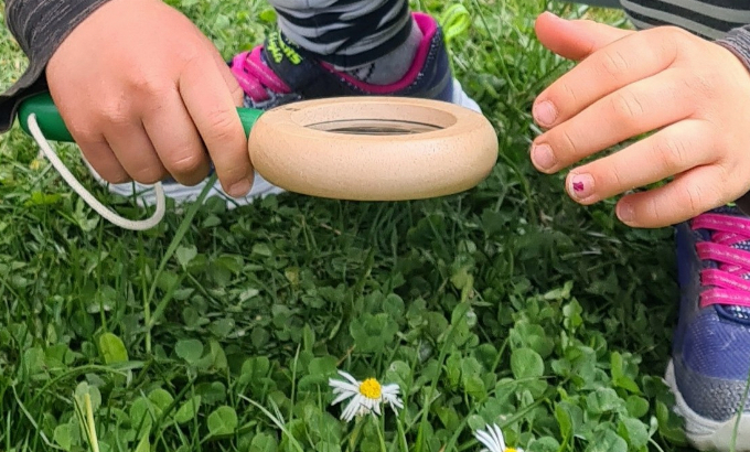 Kind hockt in der Wiese und schaut sich eine Blume durch ein Vergrößerungsglas an 