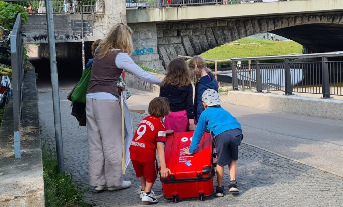 Vier Kinder und eine erwachsene Person schieben den Koffer auf einem Gehweg; die Personen sind von hinten zu sehen